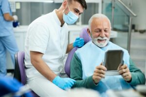 Satisfied senior man looking his teeth in a mirror after dental procedure at dentist's office.